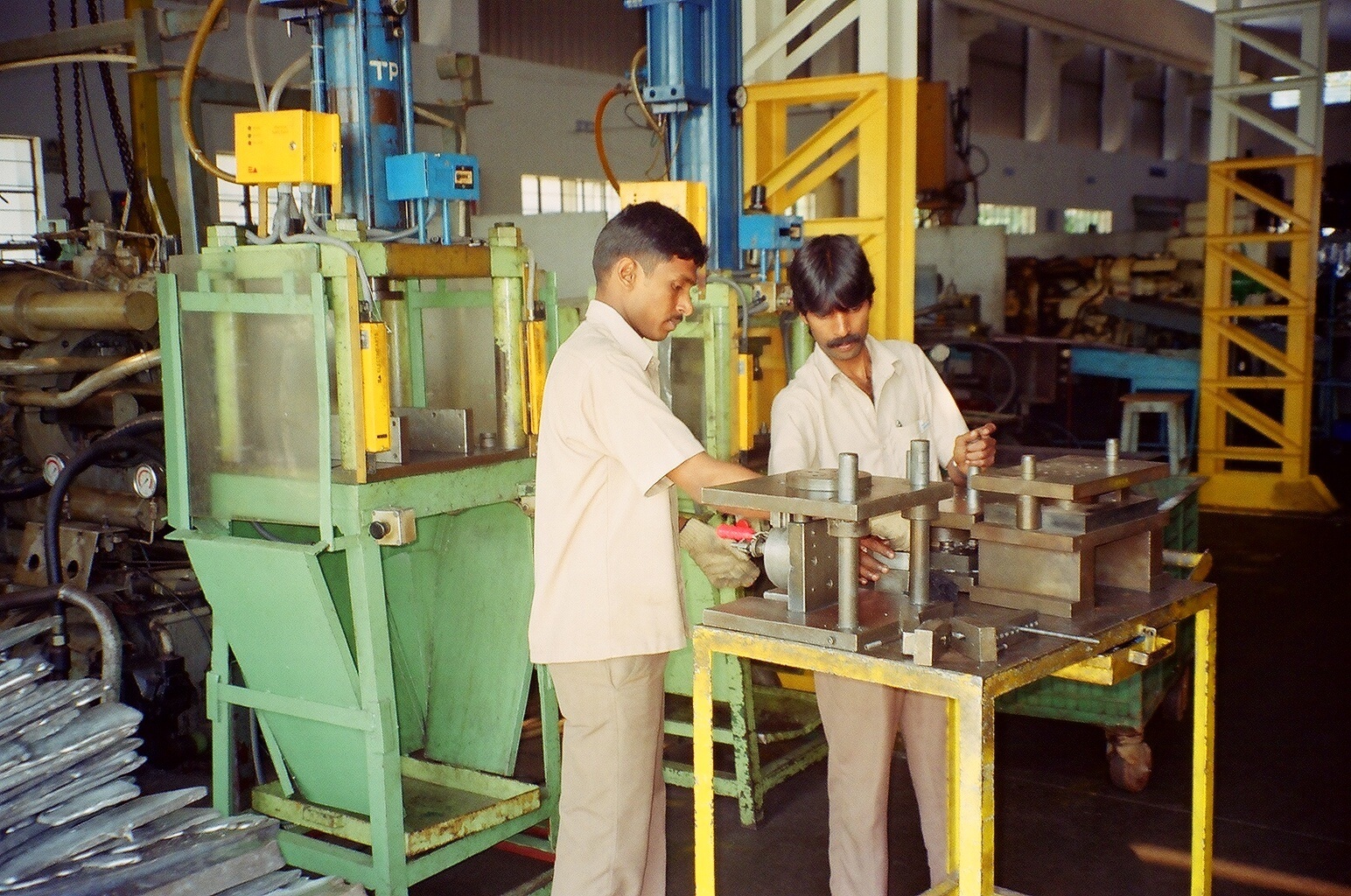 Two operators working on a die at a bench between hydraulic presses on a shop floor, finished parts stacked beside them.